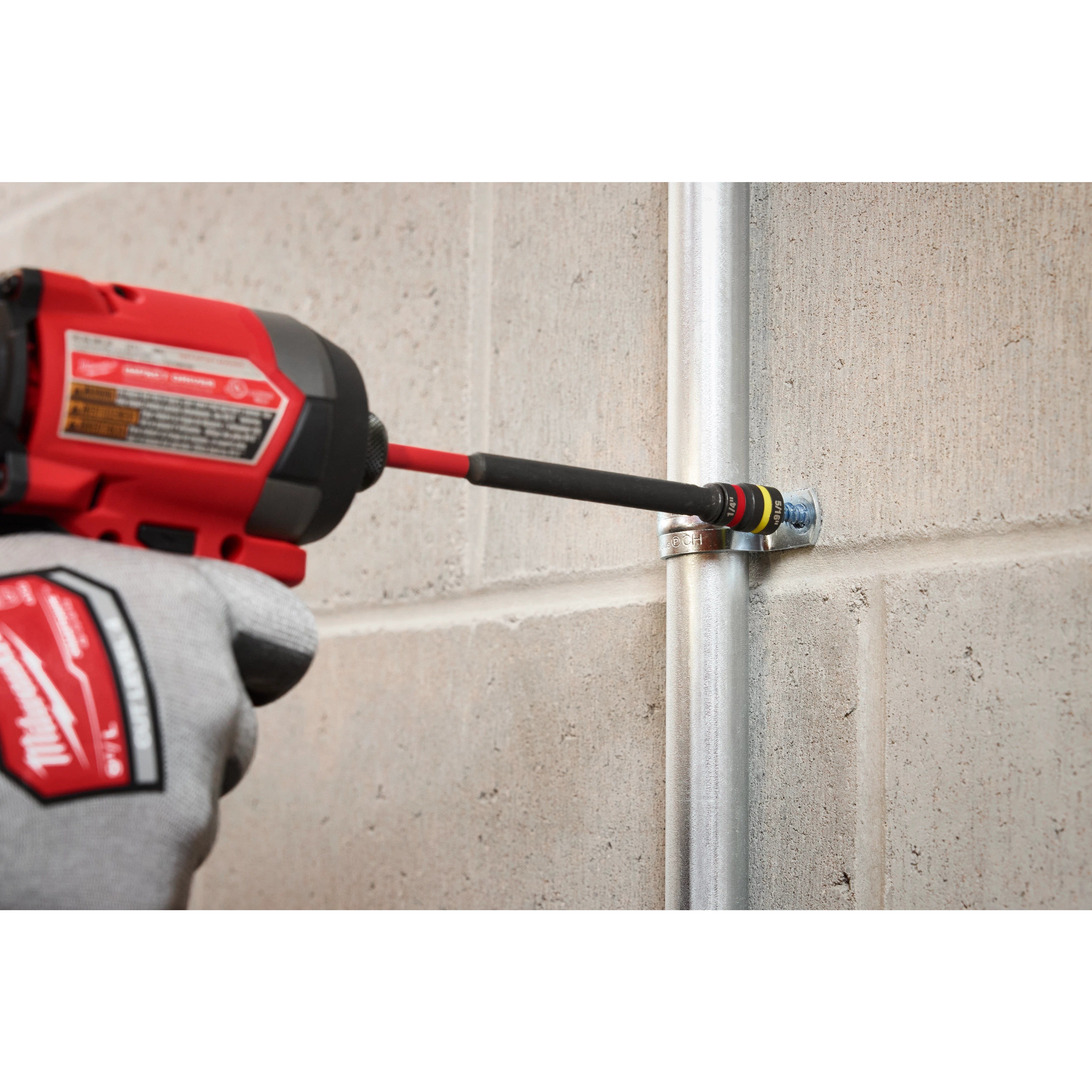 A person uses the SHOCKWAVE™ Impact Duty 1/4” and 5/16” x 6” QUIK-CLEAR™ 2-in-1 Magnetic Nut Driver to fasten a bolt onto a pipe attached to a concrete wall. The product, held in a red and black power tool, features magnetic tips for efficient and secure fastening.