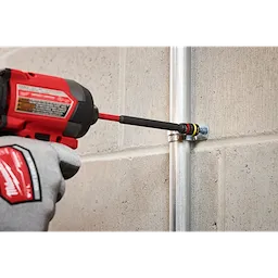 A person uses the SHOCKWAVE™ Impact Duty 1/4” and 5/16” x 6” QUIK-CLEAR™ 2-in-1 Magnetic Nut Driver to fasten a bolt onto a pipe attached to a concrete wall. The product, held in a red and black power tool, features magnetic tips for efficient and secure fastening.