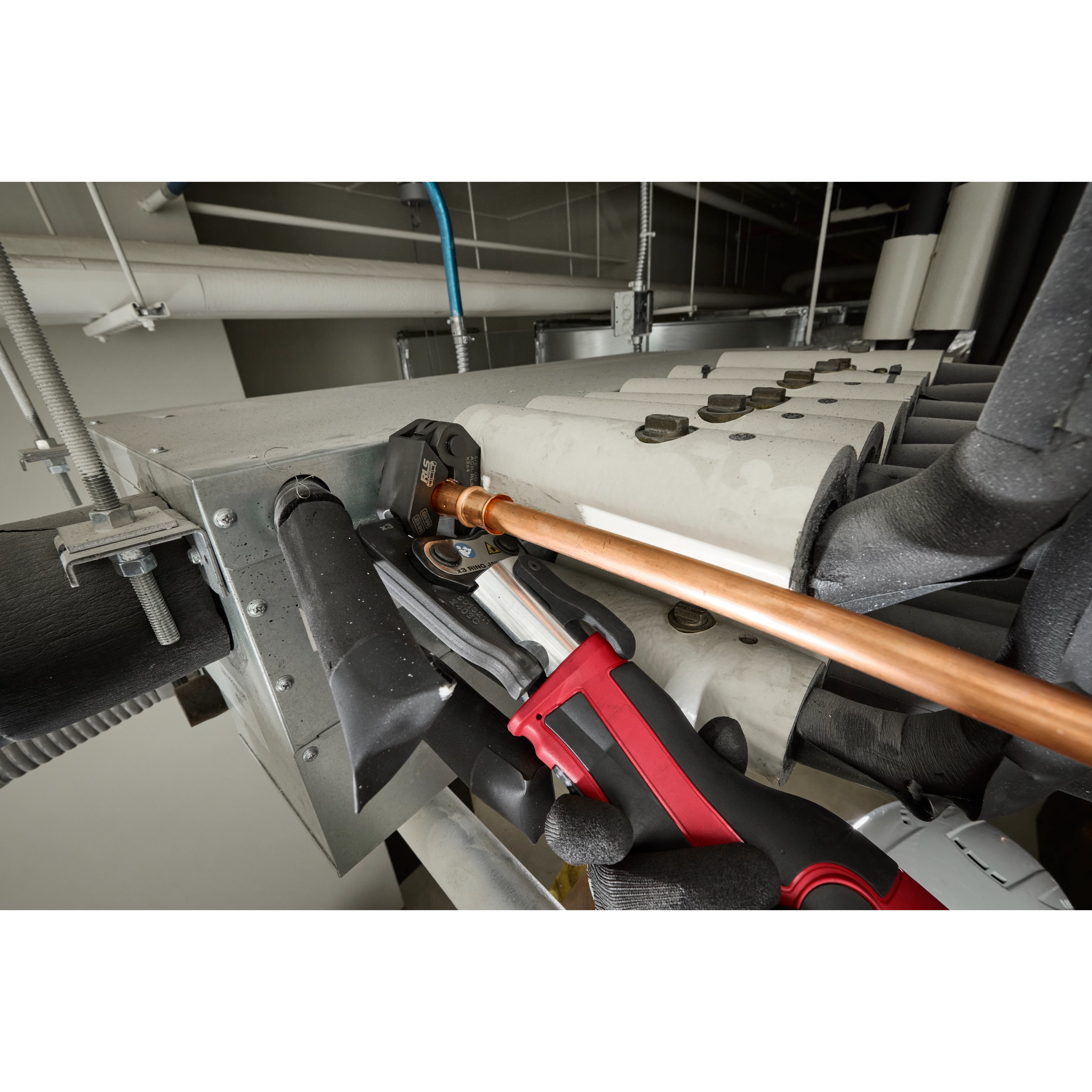 The 1/4" - 7/8" RLS® Pivoting Press Ring Kit for M12™ FORCE LOGIC™ Press Tool is being used on a copper pipe. A gloved hand operates the press tool, securing the pipe to a metal unit. Nearby black insulation and metal brackets are visible in the background.
