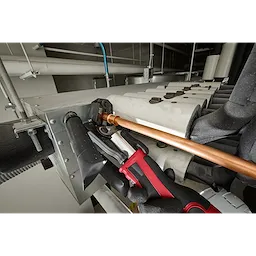 The 1/4" - 7/8" RLS® Pivoting Press Ring Kit for M12™ FORCE LOGIC™ Press Tool is being used on a copper pipe. A gloved hand operates the press tool, securing the pipe to a metal unit. Nearby black insulation and metal brackets are visible in the background.