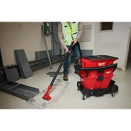 A worker uses the NEXUS™ Dedicated Debris Separator to clean debris on a construction site. The red and black machine has hoses attached and is mounted on wheels for easy mobility. The worker wears a reflective vest and operates the vacuum with a wide nozzle attachment.