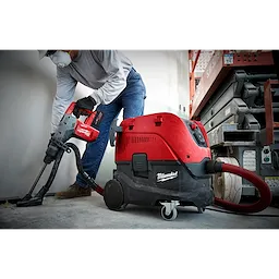 A person operates the M18 FUEL™ Rotary Hammer with Chisel Boot, M18 FUEL 1-9/16 SDS MAX Hammer Drill. The tool is red and black and is connected to a Milwaukee vacuum system for dust collection. The person wears safety gear and stands next to a construction lift.