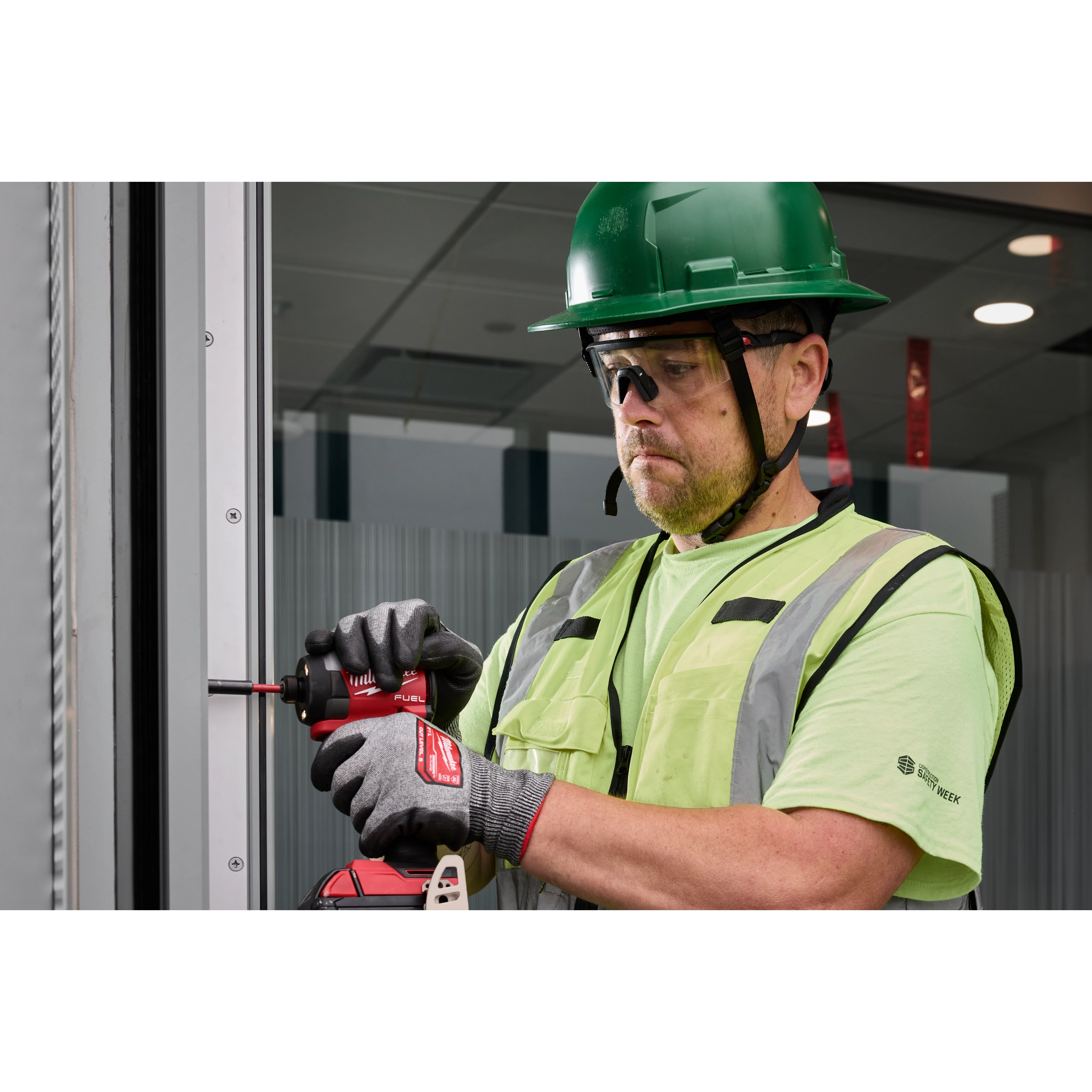 A person wearing a green safety helmet, high-visibility vest, and gloves is using a power drill to work on a metal frame. The person is also wearing Wrap Around Safety Glasses with Clear Anti-Scratch Lenses.