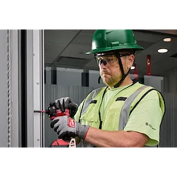 A person wearing a green safety helmet, high-visibility vest, and gloves is using a power drill to work on a metal frame. The person is also wearing Wrap Around Safety Glasses with Clear Anti-Scratch Lenses.