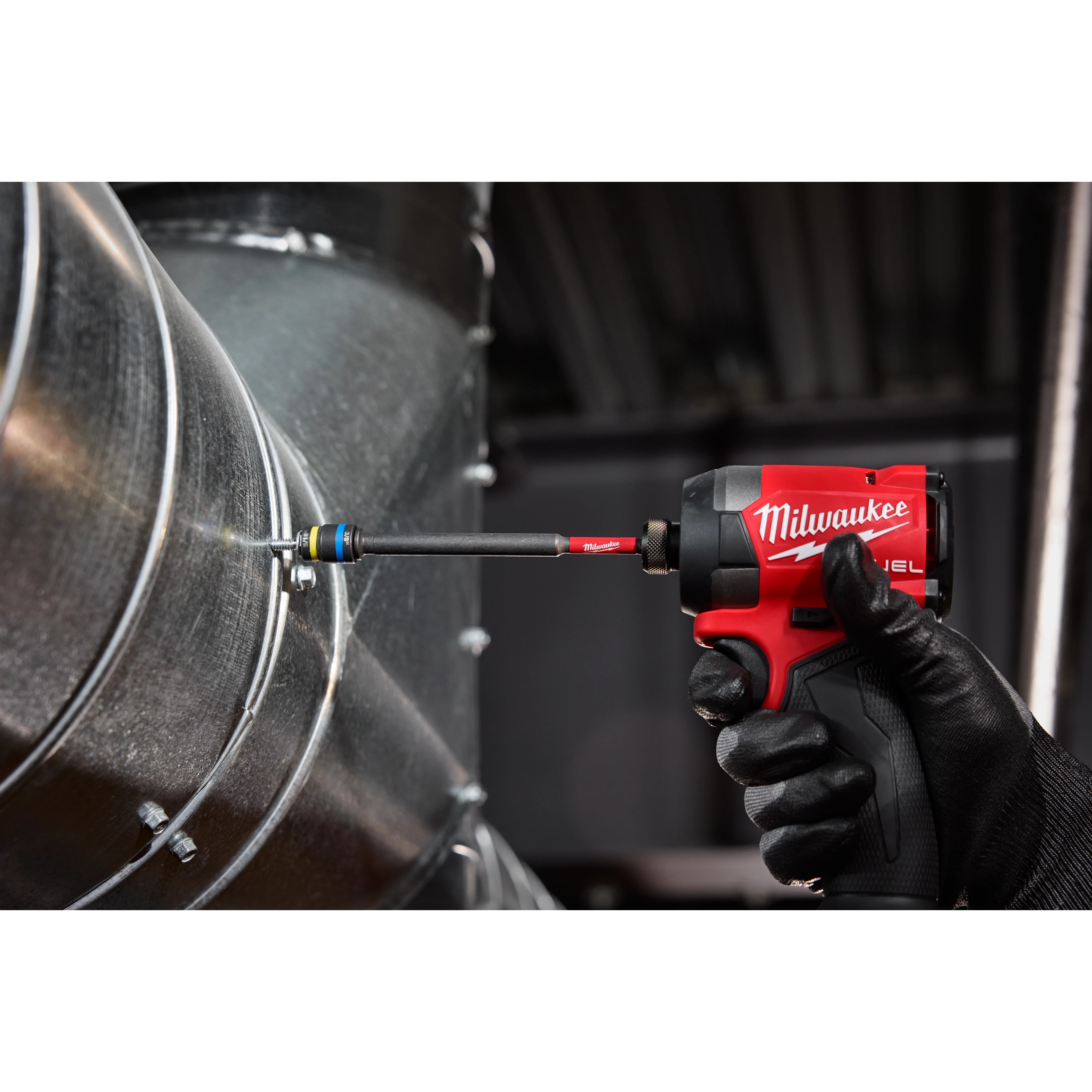 A black-gloved hand is using a red Milwaukee impact driver equipped with the SHOCKWAVE™ Impact Duty 5/16” and 3/8” x 6” QUIK-CLEAR™ 2-in-1 Magnetic Nut Driver to fasten a screw into a metallic surface. The nut driver is attached to a large cylindrical duct.