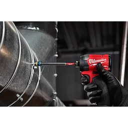 A black-gloved hand is using a red Milwaukee impact driver equipped with the SHOCKWAVE™ Impact Duty 5/16” and 3/8” x 6” QUIK-CLEAR™ 2-in-1 Magnetic Nut Driver to fasten a screw into a metallic surface. The nut driver is attached to a large cylindrical duct.