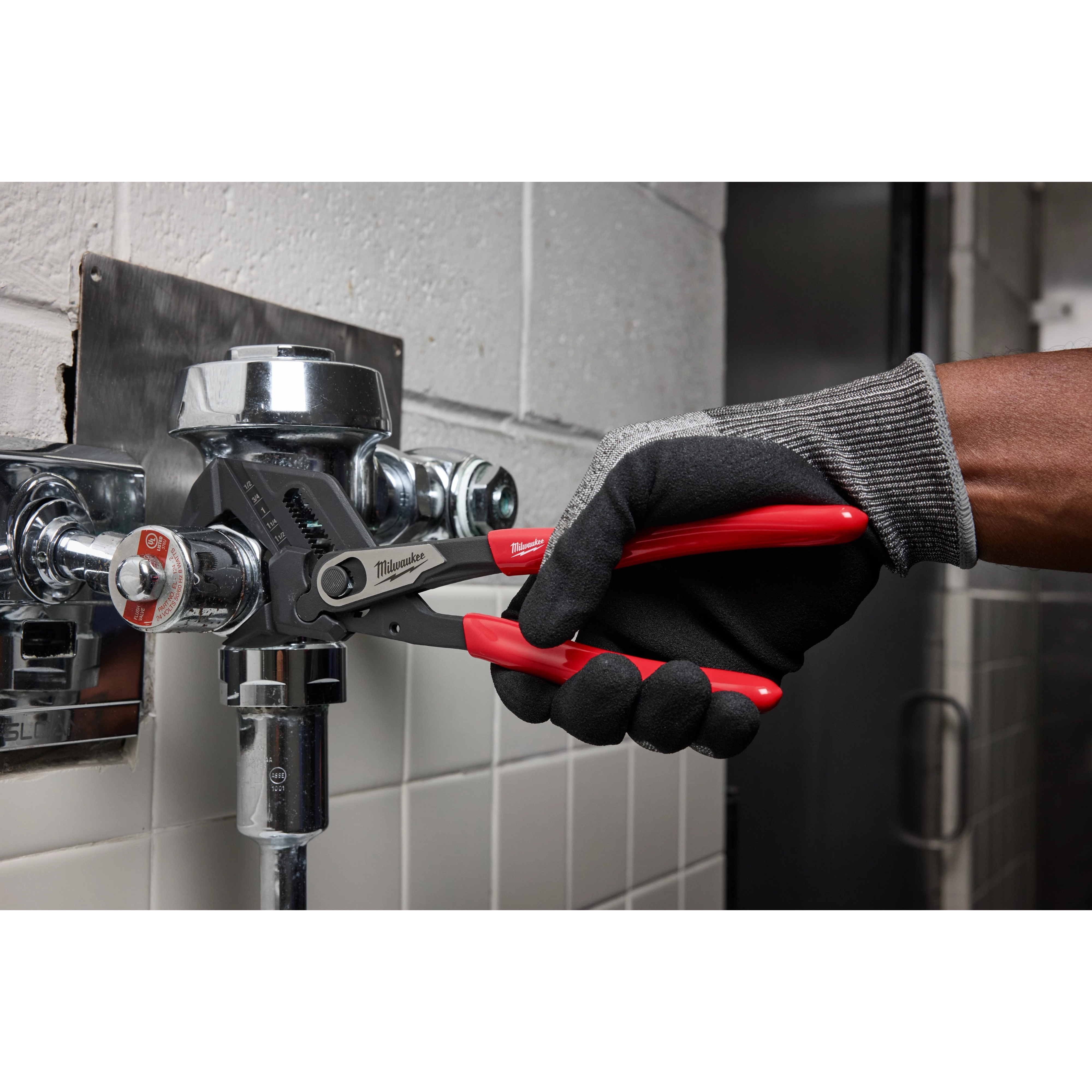 A person wearing gloves uses the 10" Pliers Wrench to tighten a metal pipe fixture in a tiled bathroom. The wrench has black jaws and red handles.