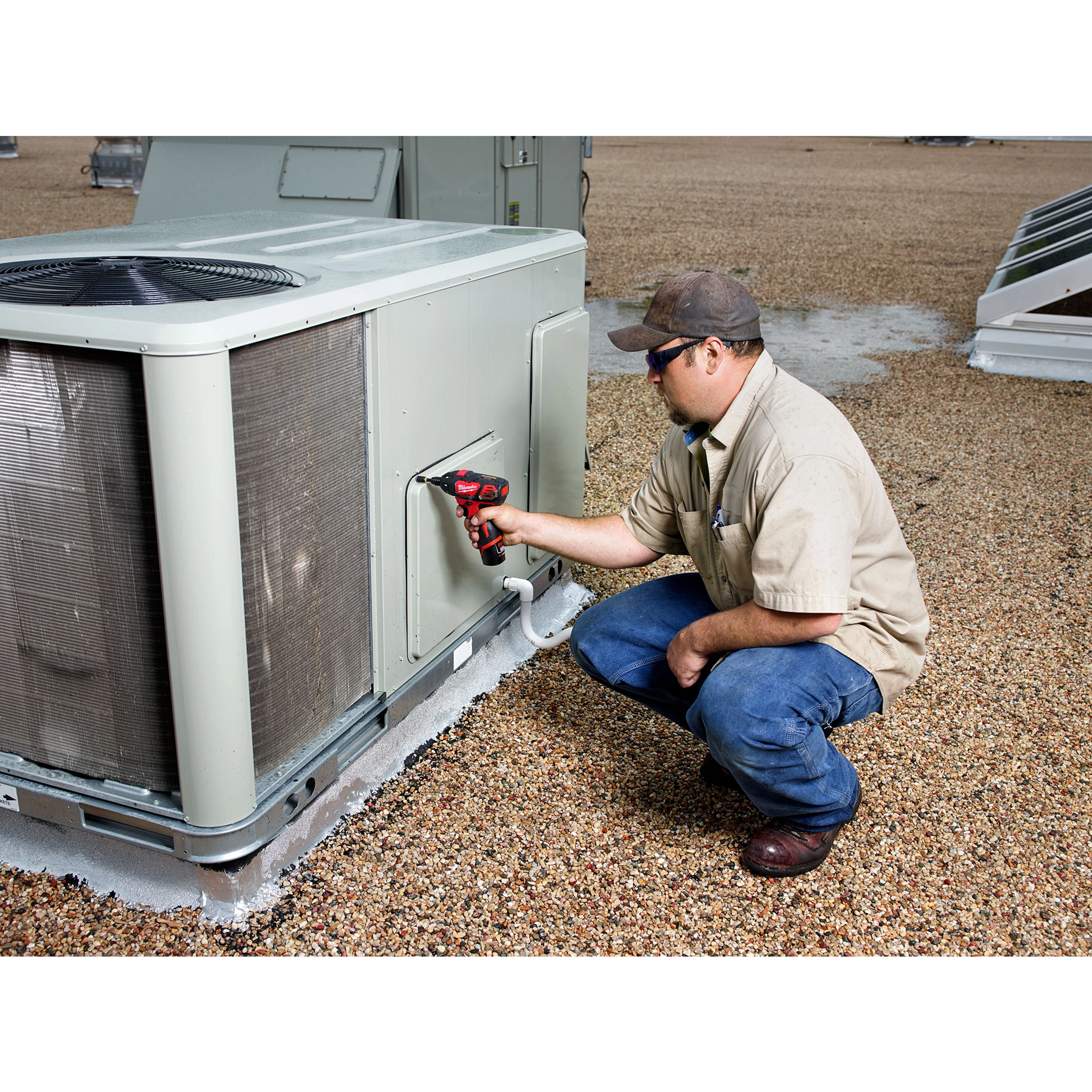 A person using an M12 Screwdriver to work on a large air conditioning unit on a gravel rooftop.
