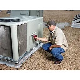 A person using an M12 Screwdriver to work on a large air conditioning unit on a gravel rooftop.