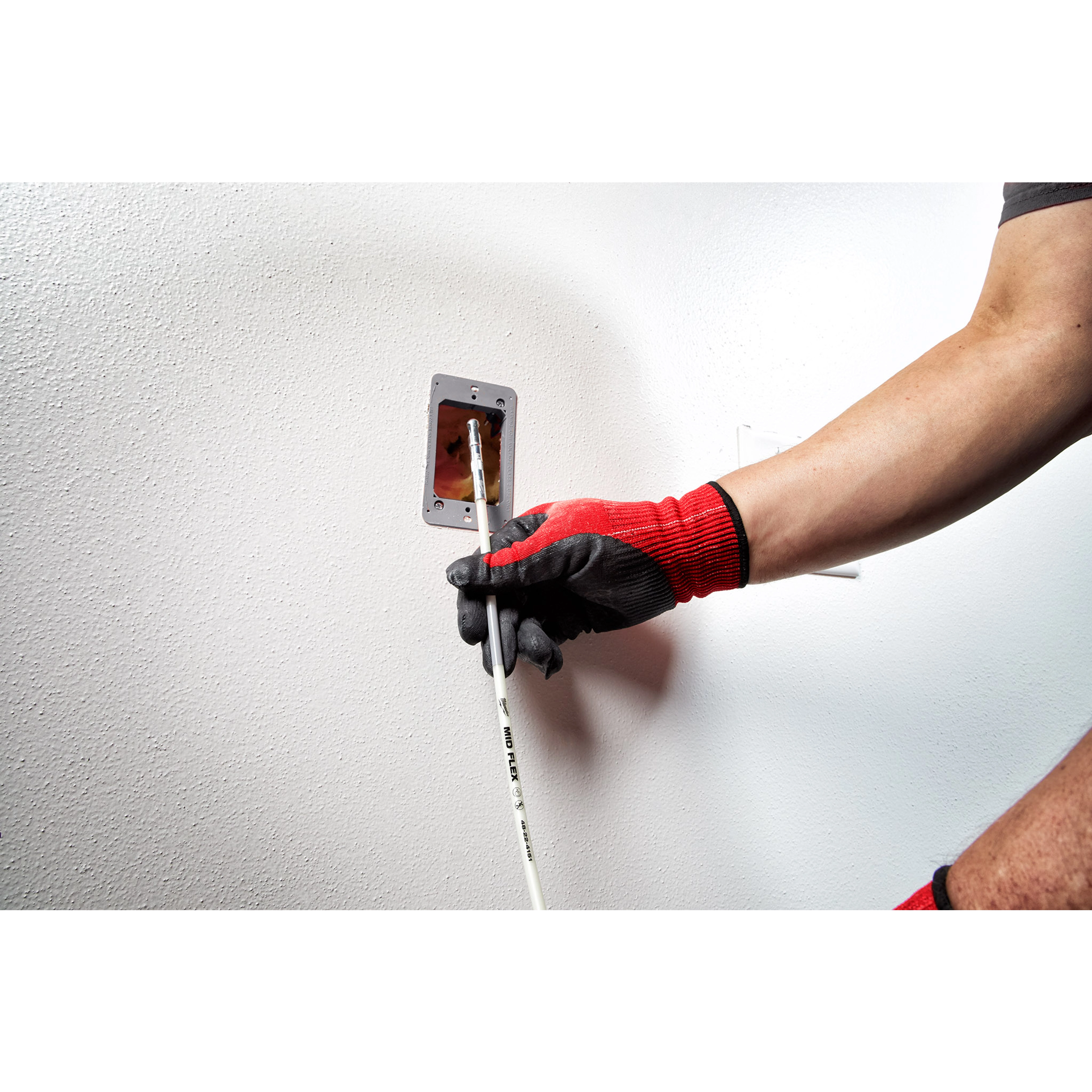 A person wearing red and black gloves uses the 5' Mid Flex Fish Stick to pull wires through an electrical outlet box in a wall.