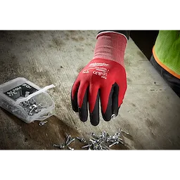 A person wearing Milwaukee Cut 1 Dipped Gloves is picking up screws from a wooden surface next to an open plastic container filled with screws. The gloves are red with black fingertips and have protective labels on the back.