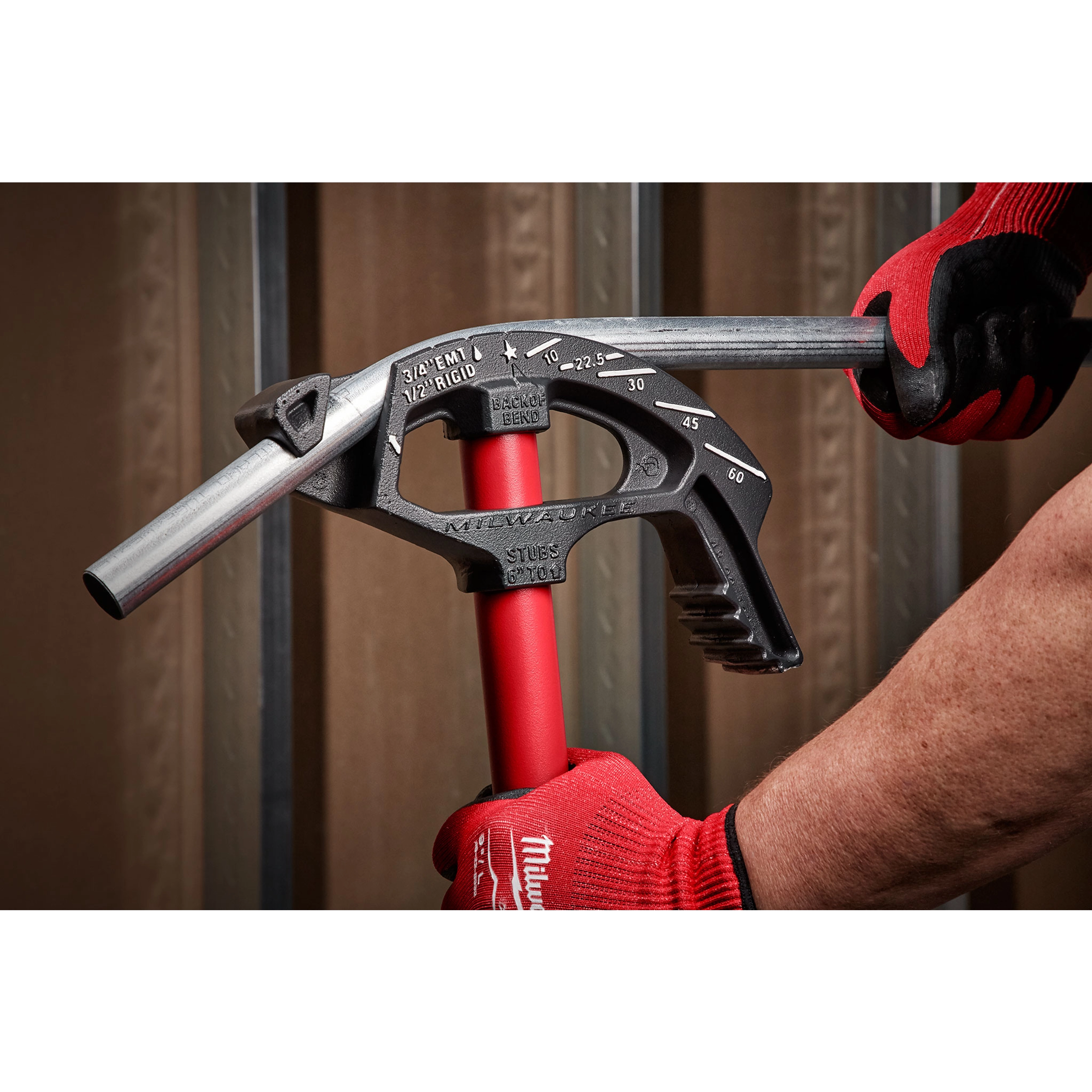 A person wearing red gloves uses a 3/4" EMT Iron Conduit Bender to bend a metal conduit. The bender features angle marks and a red handle.