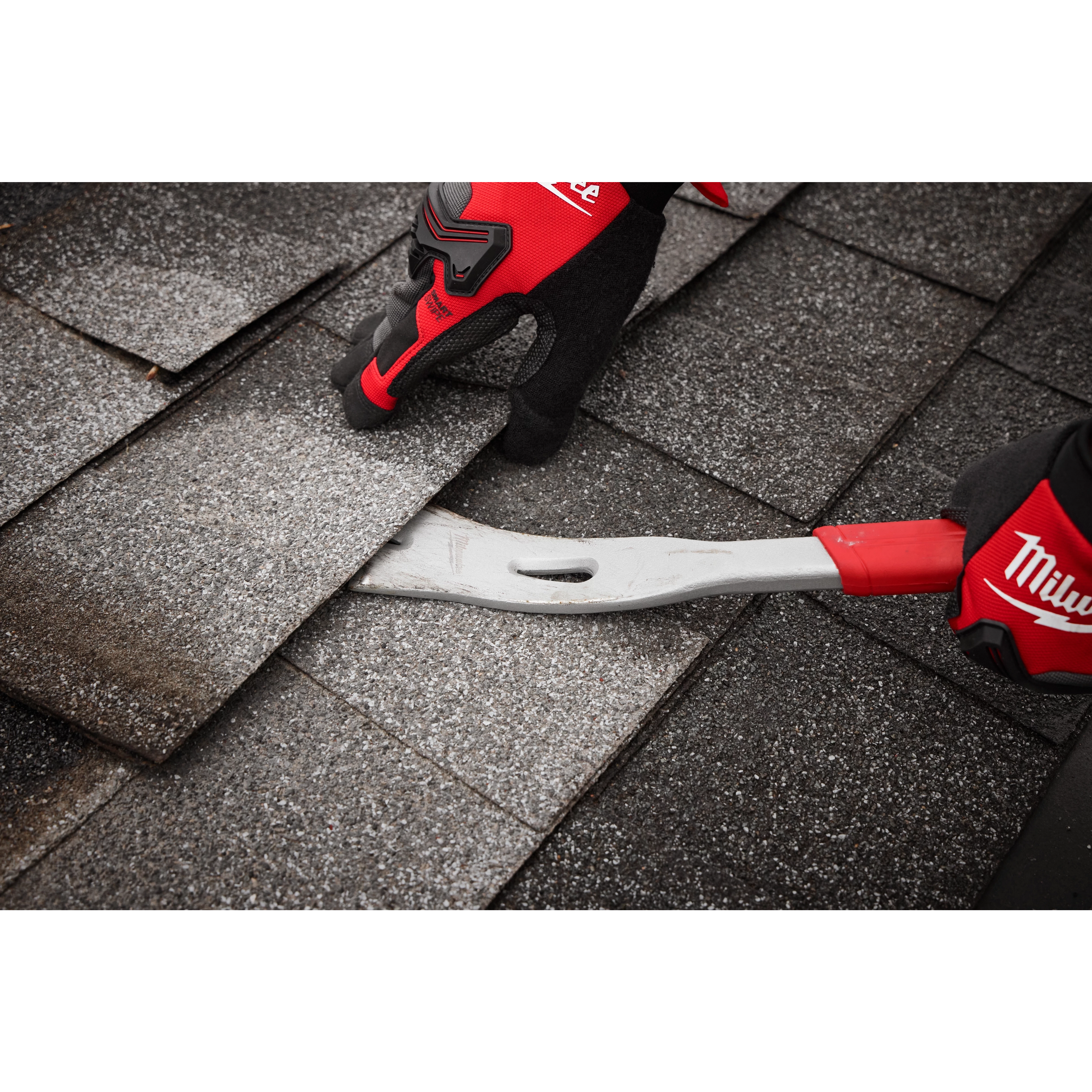 The 16" Molding Puller from Milwaukee is being used to remove roof shingles. A person wearing protective gloves grips the metal tool with a red handle as they lift the edge of the shingles, revealing the layer beneath. There are multiple shingles visible in the background.