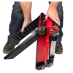 Hands unfolding the legs of a red, compact M18 Rocket Light placed on the floor. The person is wearing blue jeans and brown boots.