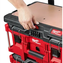 A hand grips the handle of a stacked red and black PACKOUT Customizable Work Top with a wooden surface and metal latches.