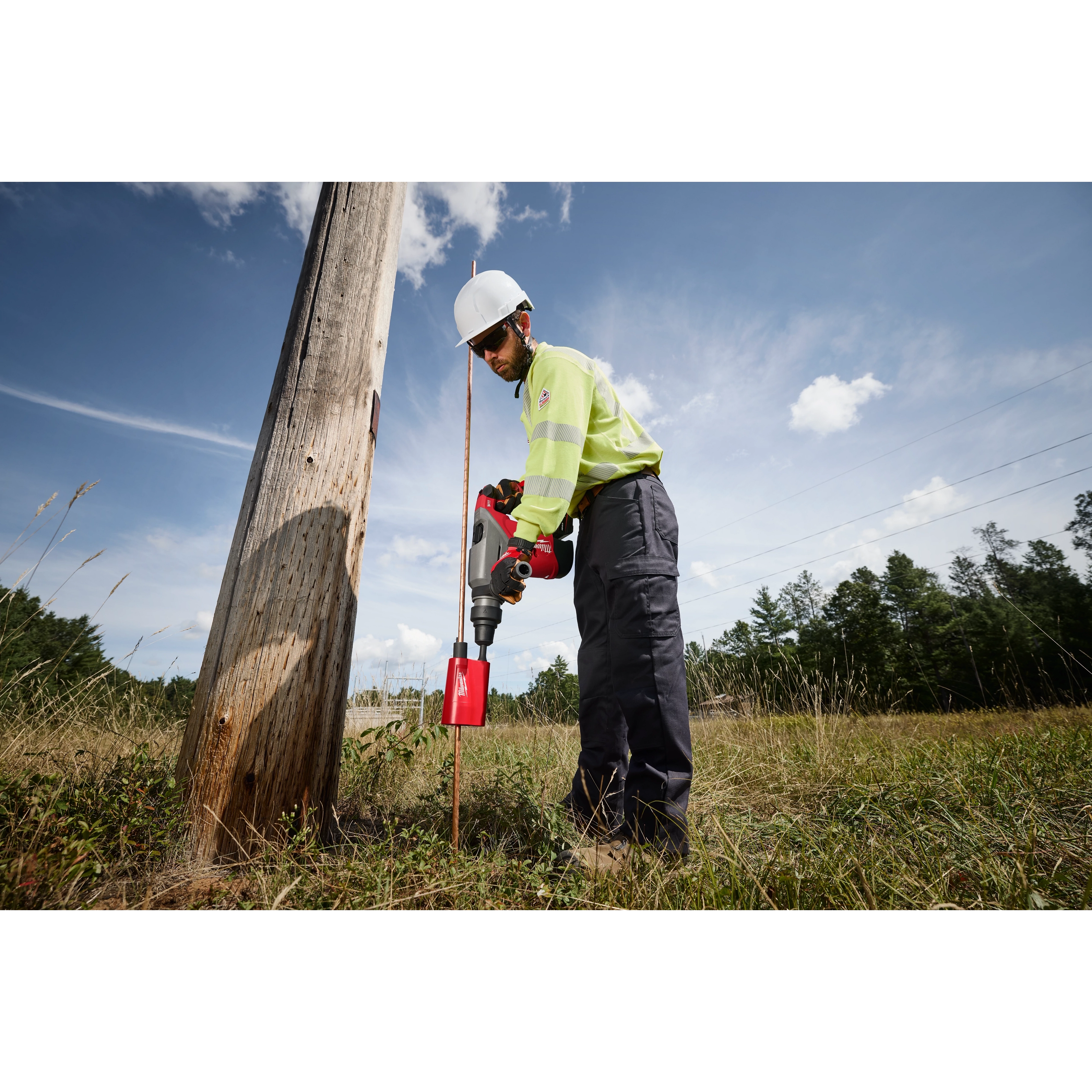 A construction worker uses the Milwaukee SDS Max Side Load Ground Rod Driver to drive a grounding rod into the earth next to a wooden utility pole in an open field.