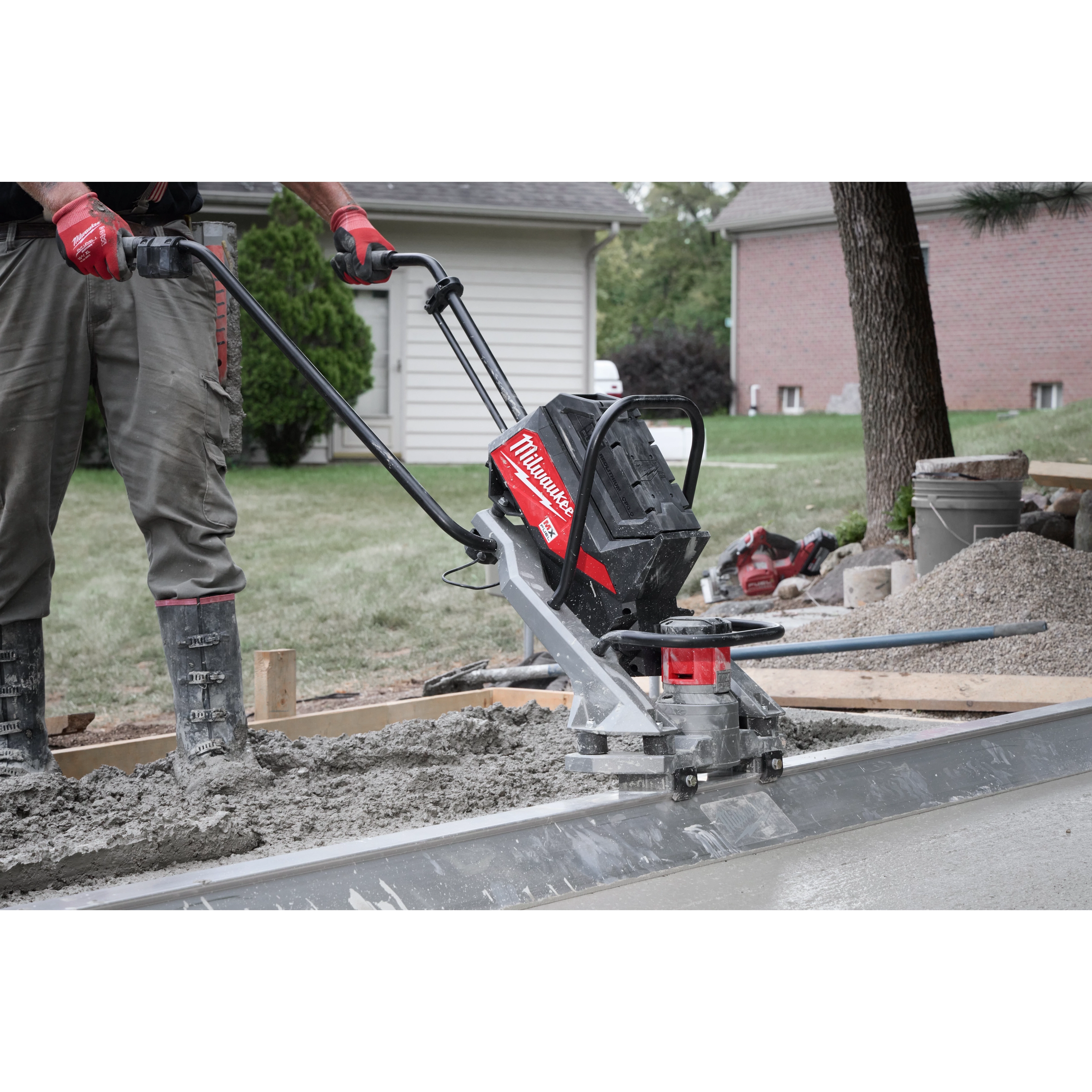The image shows a construction worker using a Milwaukee MX FUEL™ Vibratory Screed to level wet concrete on a site. The worker grips the machine's handle, guiding it over the surface. Nearby are piles of gravel and concrete tools with residential houses in the background.