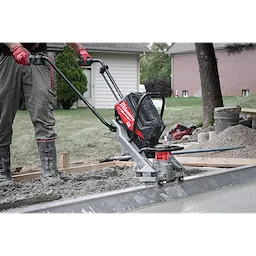 The image shows a construction worker using a Milwaukee MX FUEL™ Vibratory Screed to level wet concrete on a site. The worker grips the machine's handle, guiding it over the surface. Nearby are piles of gravel and concrete tools with residential houses in the background.