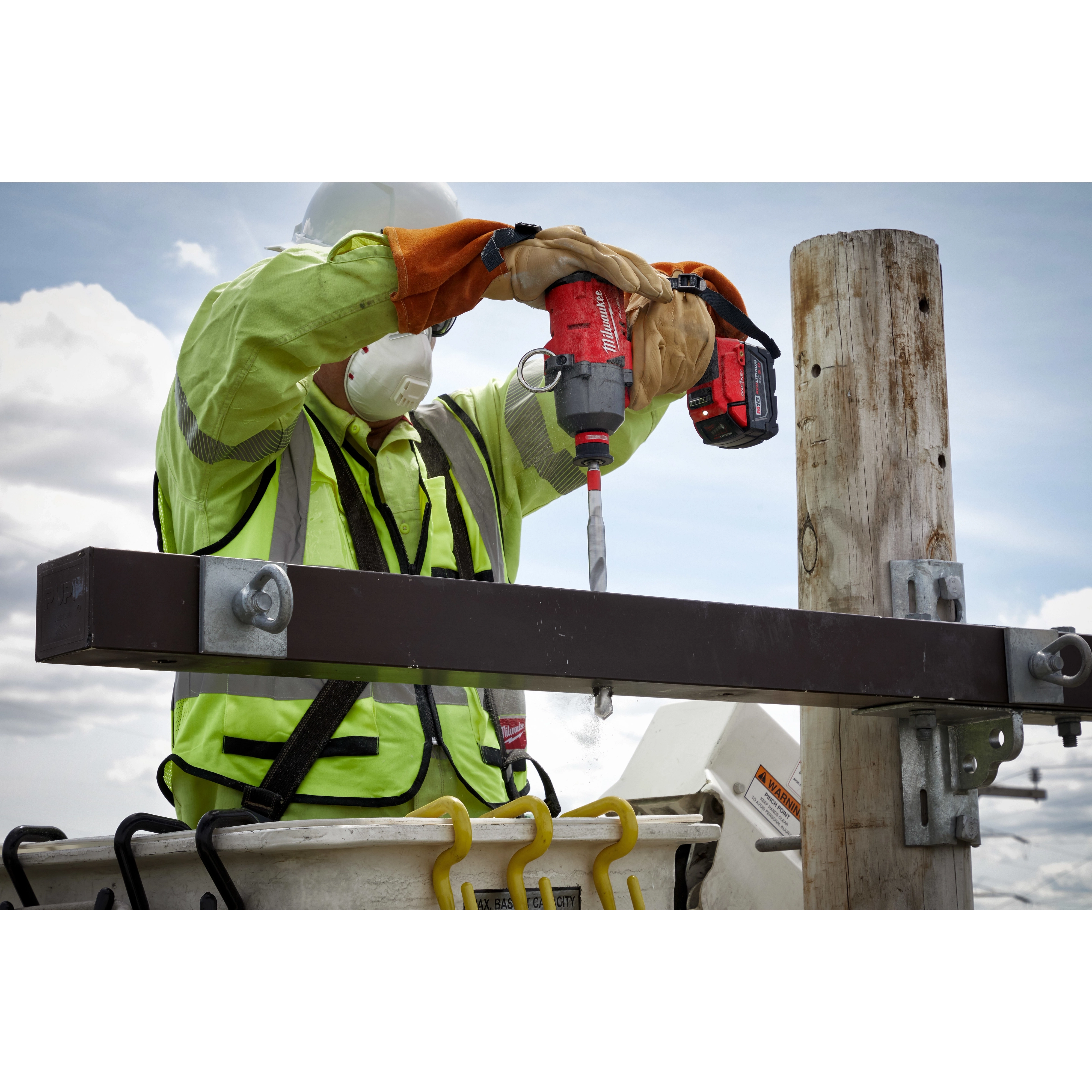 A worker in safety gear uses a red cordless drill to operate the 13/16"x12" Shockwave Lineman's Fiberglass Drill Bit on a metal beam attached to a wooden pole. The worker is standing in a bucket truck against a backdrop of sky and clouds.