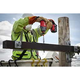 A worker in safety gear uses a red cordless drill to operate the 13/16"x12" Shockwave Lineman's Fiberglass Drill Bit on a metal beam attached to a wooden pole. The worker is standing in a bucket truck against a backdrop of sky and clouds.