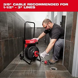 Milwaukee drain cleaning machine being used in a tiled bathroom by a person wearing gloves. The machine has a black reel and red accents, with a recommendation for a 3/8" cable suitable for 1-1/2" to 3" lines displayed above.