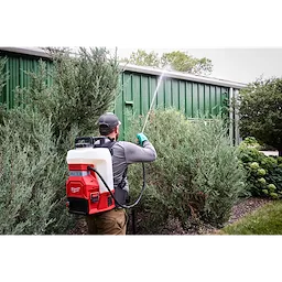 A person wearing a grey shirt and cap uses an M18™ 4-Gallon Backpack Sprayer with SWITCH TANK™ to spray liquid onto tall shrubbery beside a green metal building. The sprayer is red and white with a hose extending to the nozzle in hand.
