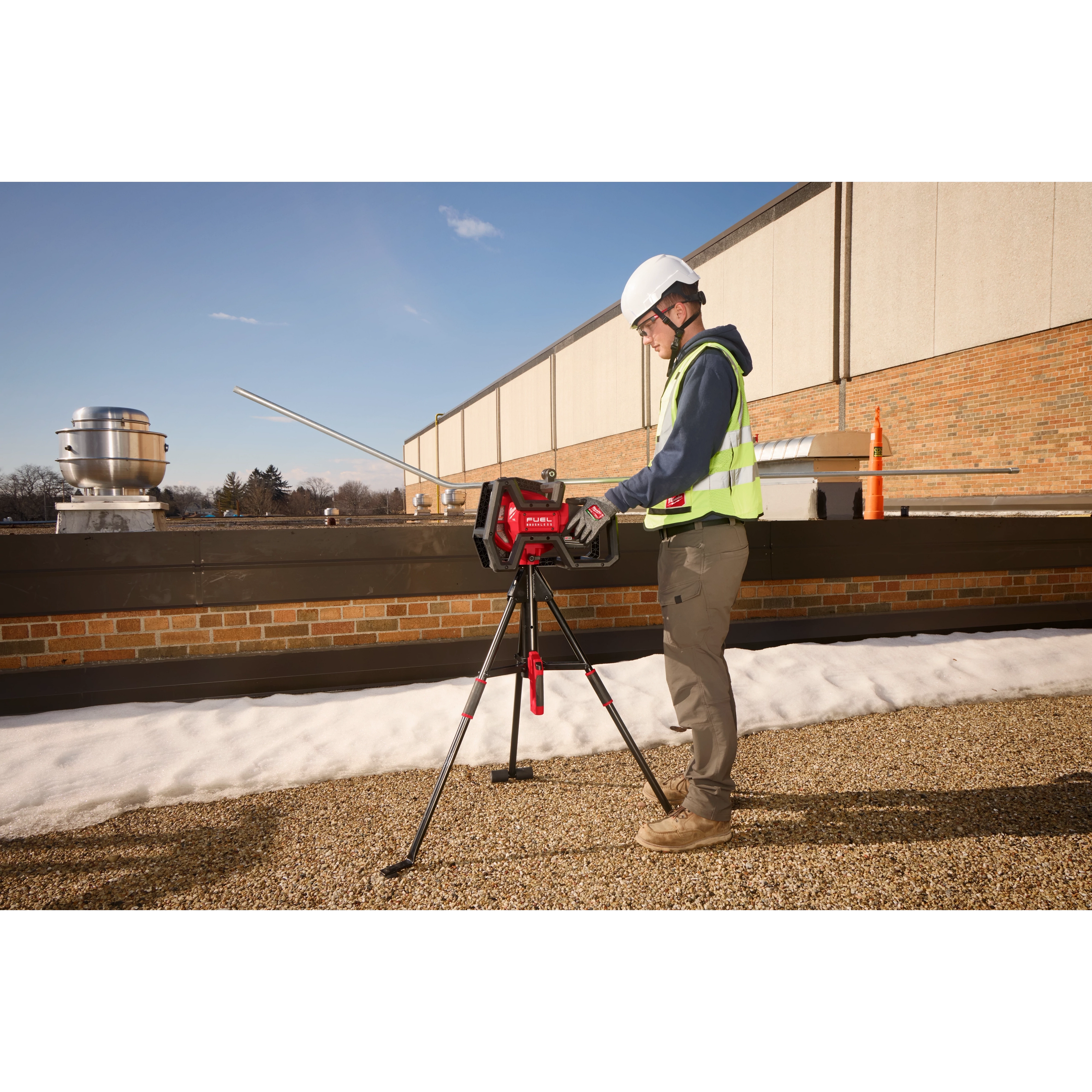 A construction worker operates the Milwaukee M18 FUEL Pipe Threader on a rooftop. The red pipe threader is mounted on a tripod, and the worker wears a hard hat and safety vest. Various pipes and equipment are visible in the background.