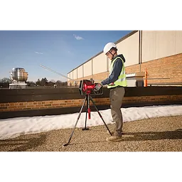 A construction worker operates the Milwaukee M18 FUEL Pipe Threader on a rooftop. The red pipe threader is mounted on a tripod, and the worker wears a hard hat and safety vest. Various pipes and equipment are visible in the background.