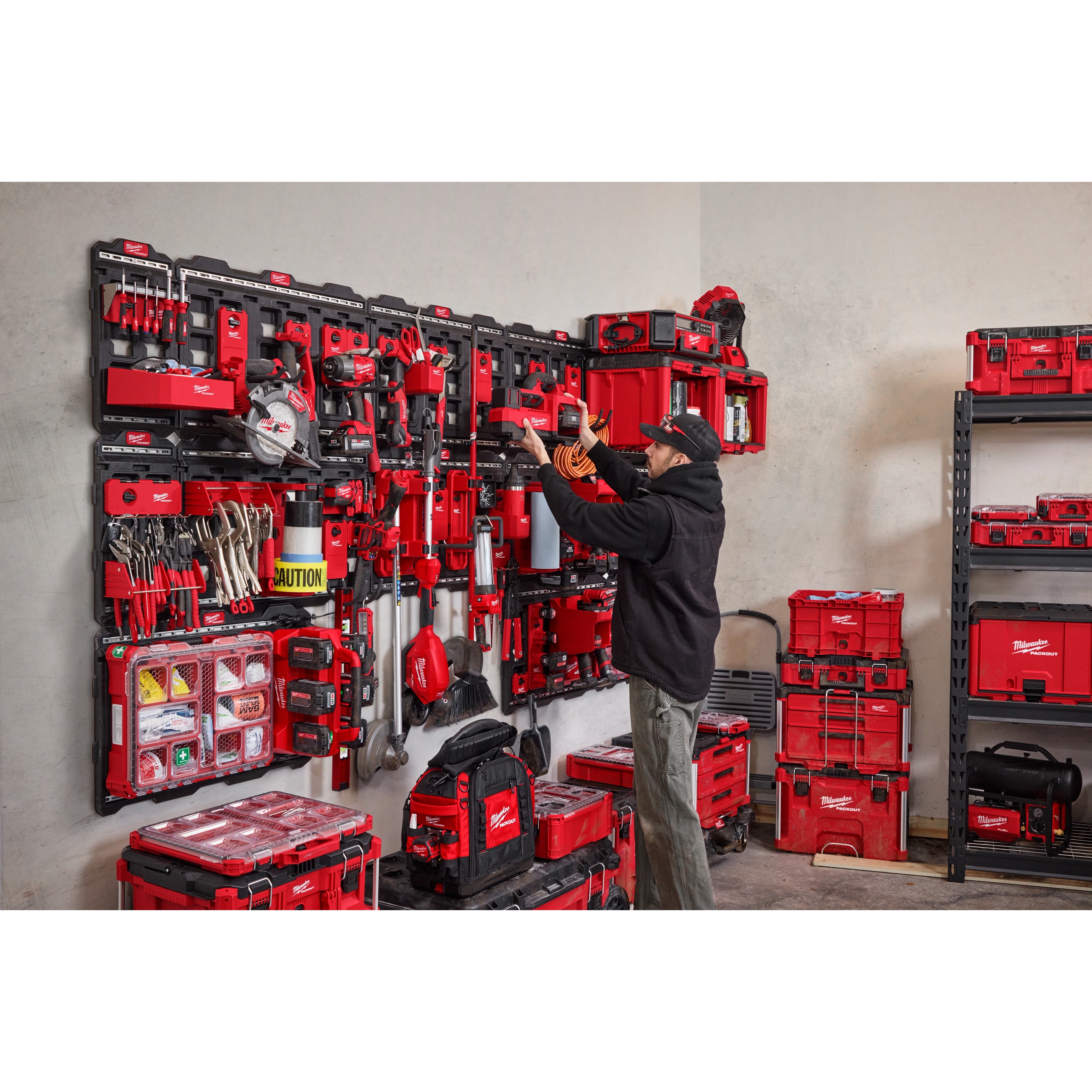 A man organizes tools using the PACKOUT™ Large Wall Plate with multiple shelves and bins. The wall plate and storage units, all in red and black, hold a variety of tools and accessories. The room has additional storage units and a shelf with more toolboxes.