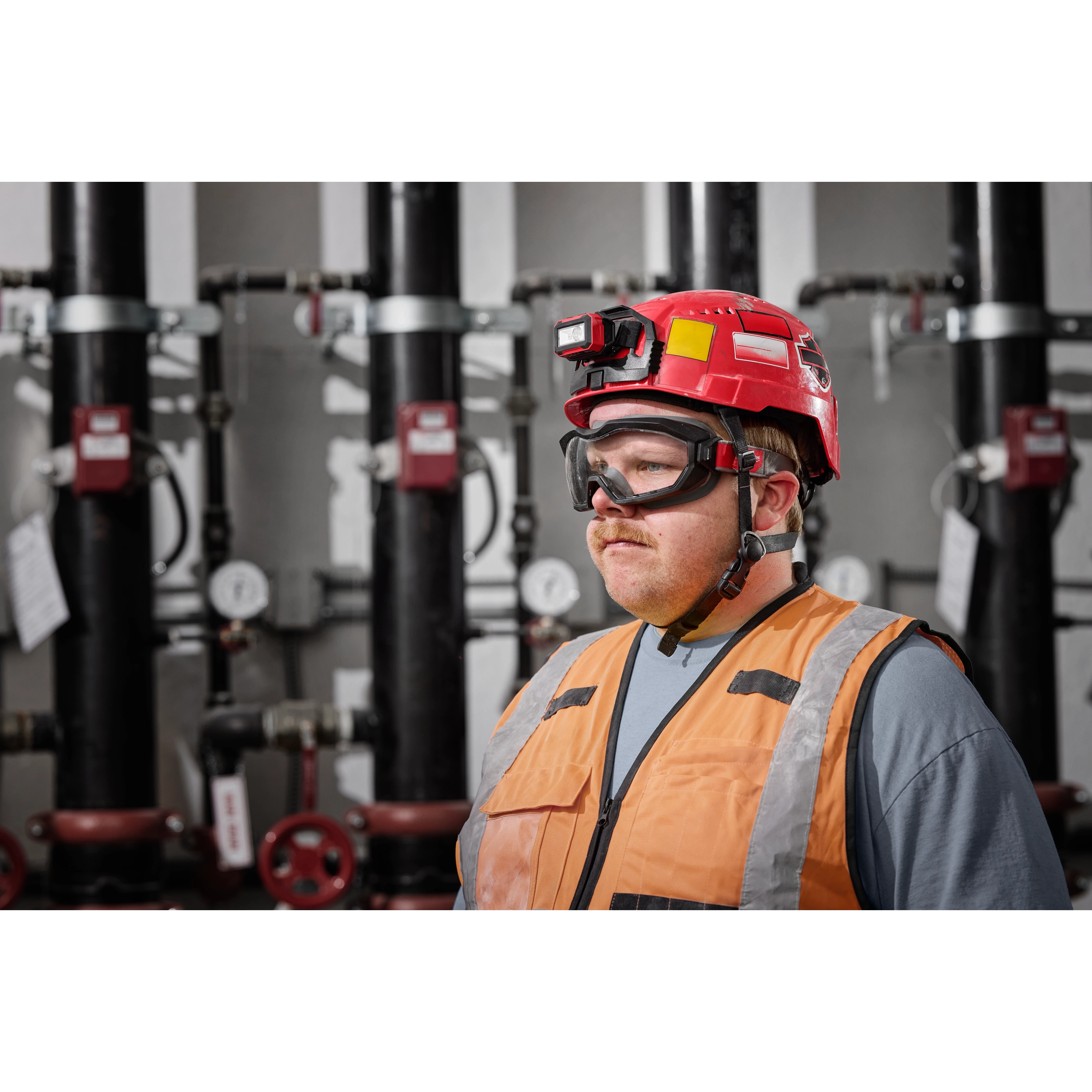 A worker wearing Non-Vented Goggles - Clear Dual Coat Lens, a red hard hat with a light, and an orange safety vest in an industrial setting.
