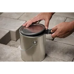 Hands using a Multi-function Compact Utility Knife to open the lid of a silver paint can. The knife is black with red accents and is being used to pry the lid open. Stone tiles are visible in the background.