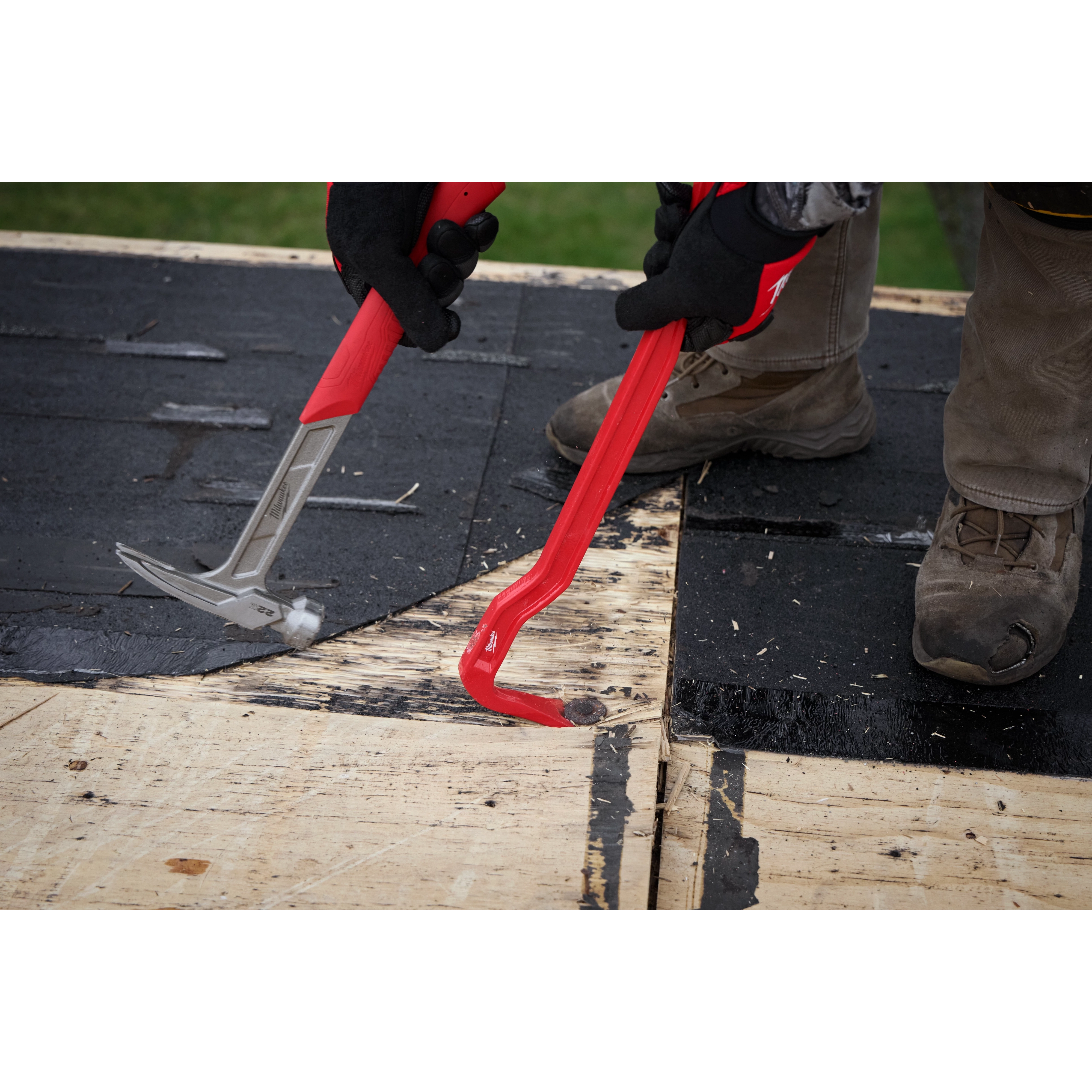 Two gloved hands holding an 18" Wrecking Bar and a roofing hammer, removing shingles from a wooden roof. The 18" Wrecking Bar is red with a flat, beveled end designed for prying. The workers wear protective clothing, including boots, and are on a partially stripped section of roofing.
