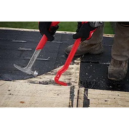 Two gloved hands holding an 18" Wrecking Bar and a roofing hammer, removing shingles from a wooden roof. The 18" Wrecking Bar is red with a flat, beveled end designed for prying. The workers wear protective clothing, including boots, and are on a partially stripped section of roofing.