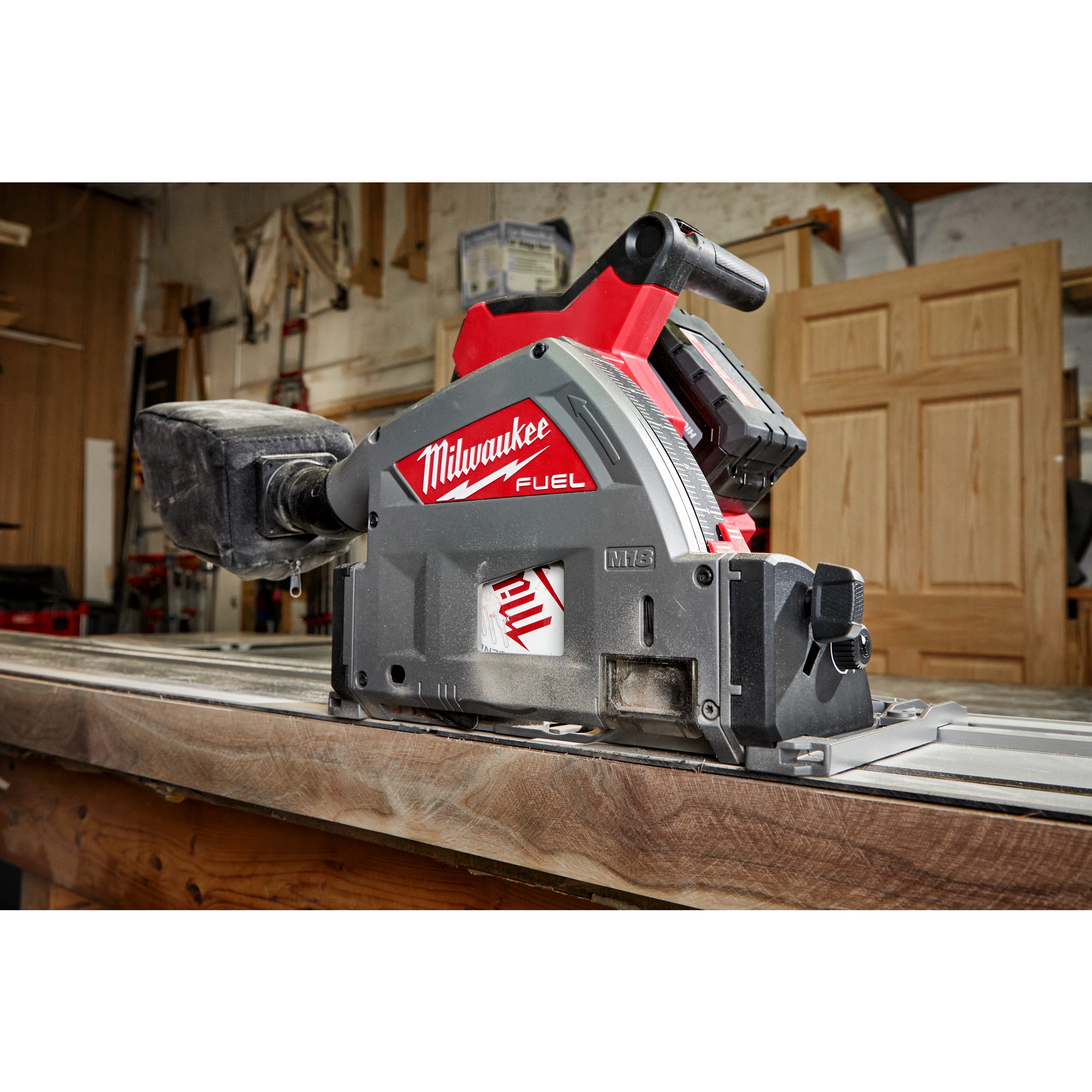 Milwaukee M18 FUEL 6 1/2" Plunge Track Saw placed on a wooden surface in a workshop with tools and doors in the background.