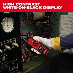 A person uses a Milwaukee clamp meter to measure electrical current inside a panel. The meter features a high-contrast white-on-black display for easy reading. The person is wearing protective gloves while handling the device.