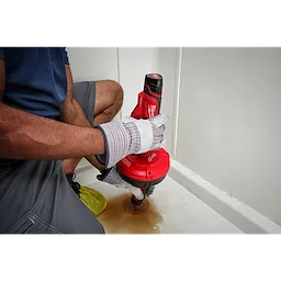 Person using a TRAPSNAKE 25' Auger on a bathroom floor, wearing safety gloves and work clothes, with a red auger machine.
