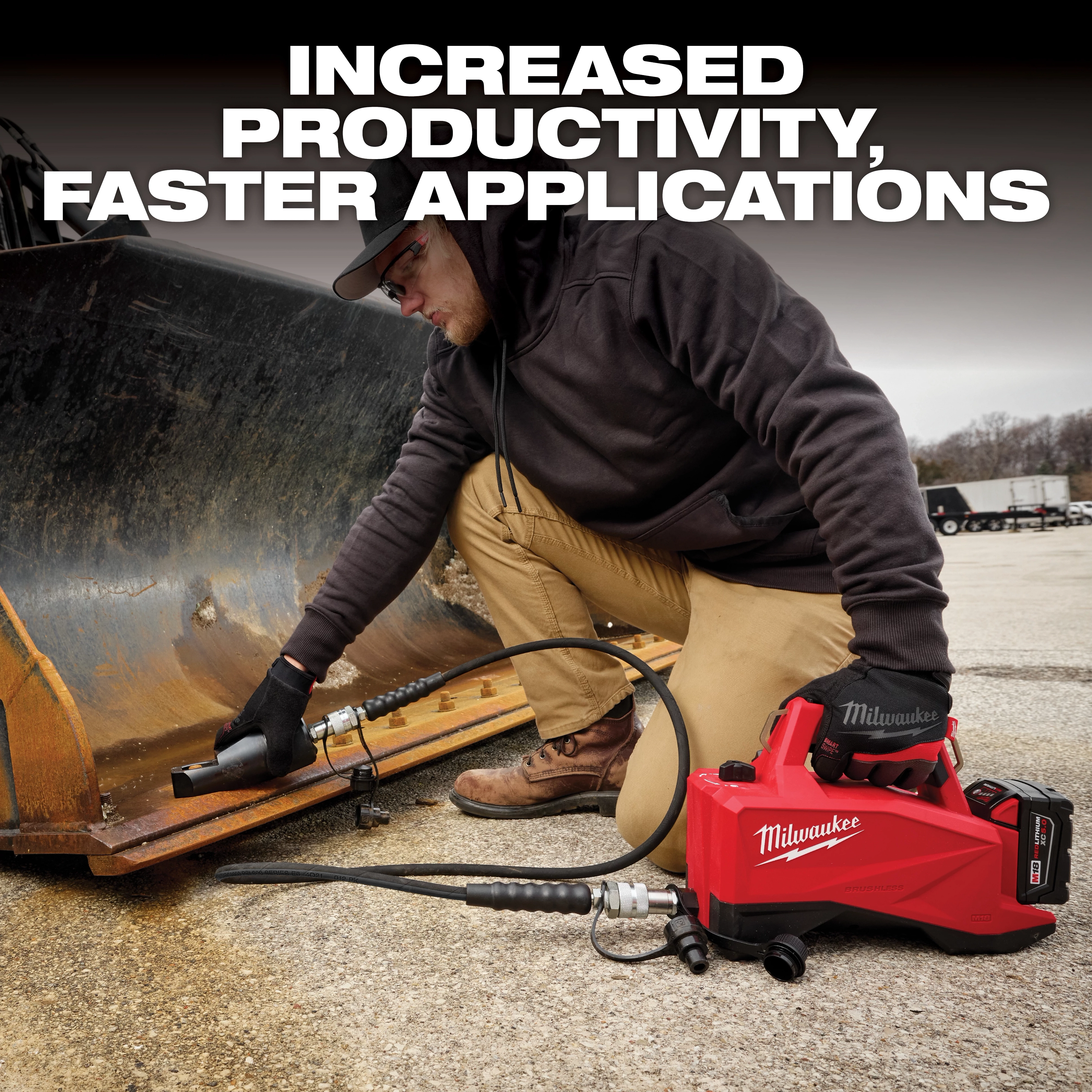 A person kneels on the ground and uses a Milwaukee hydraulic tool to repair heavy machinery. The tool is red, portable, and connected to a hose, emphasizing increased productivity and faster applications. Industrial equipment and trucks are visible in the background.