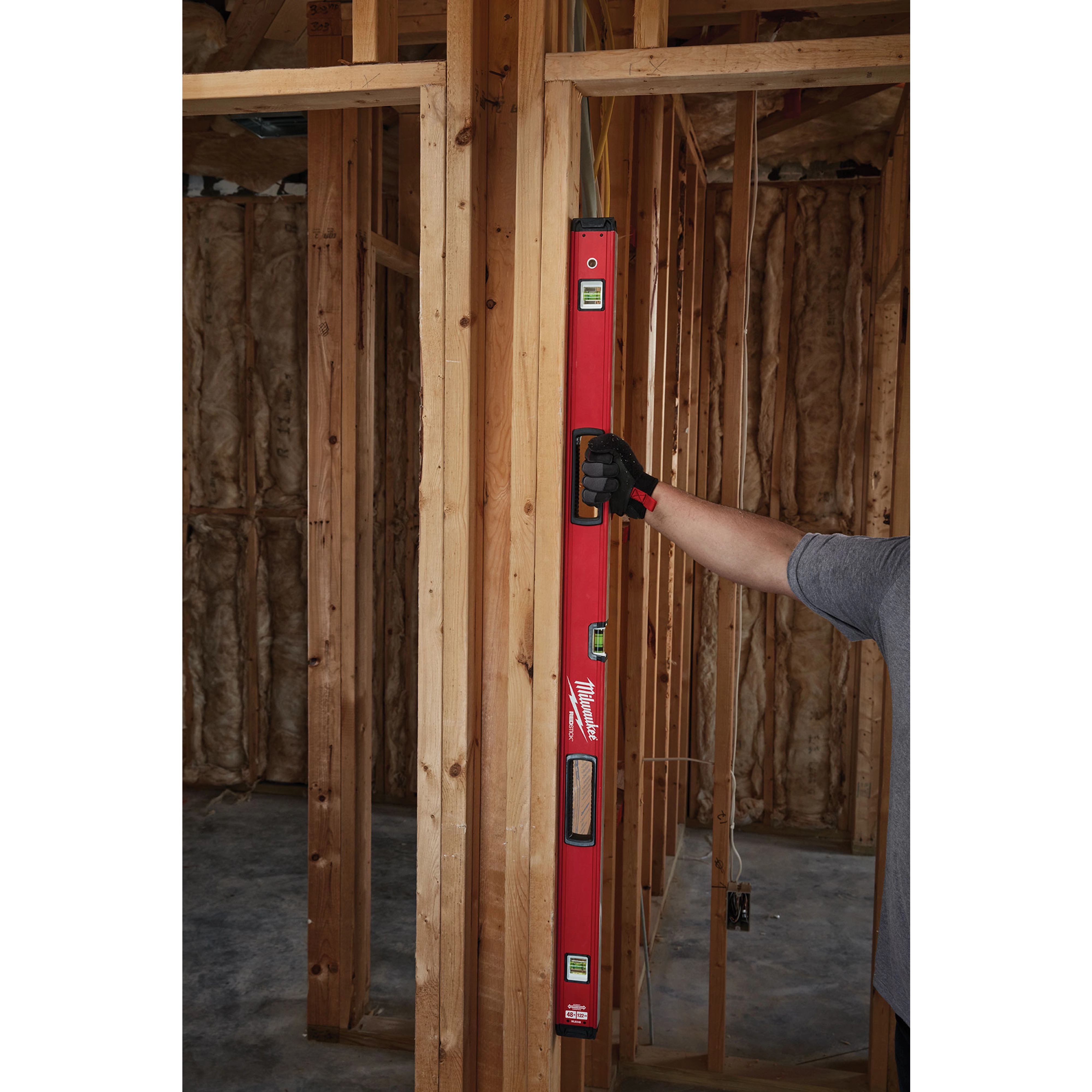 Person using a REDSTICK Level on wooden frame construction. The level has a red body, three spirit vials, and is held vertically.