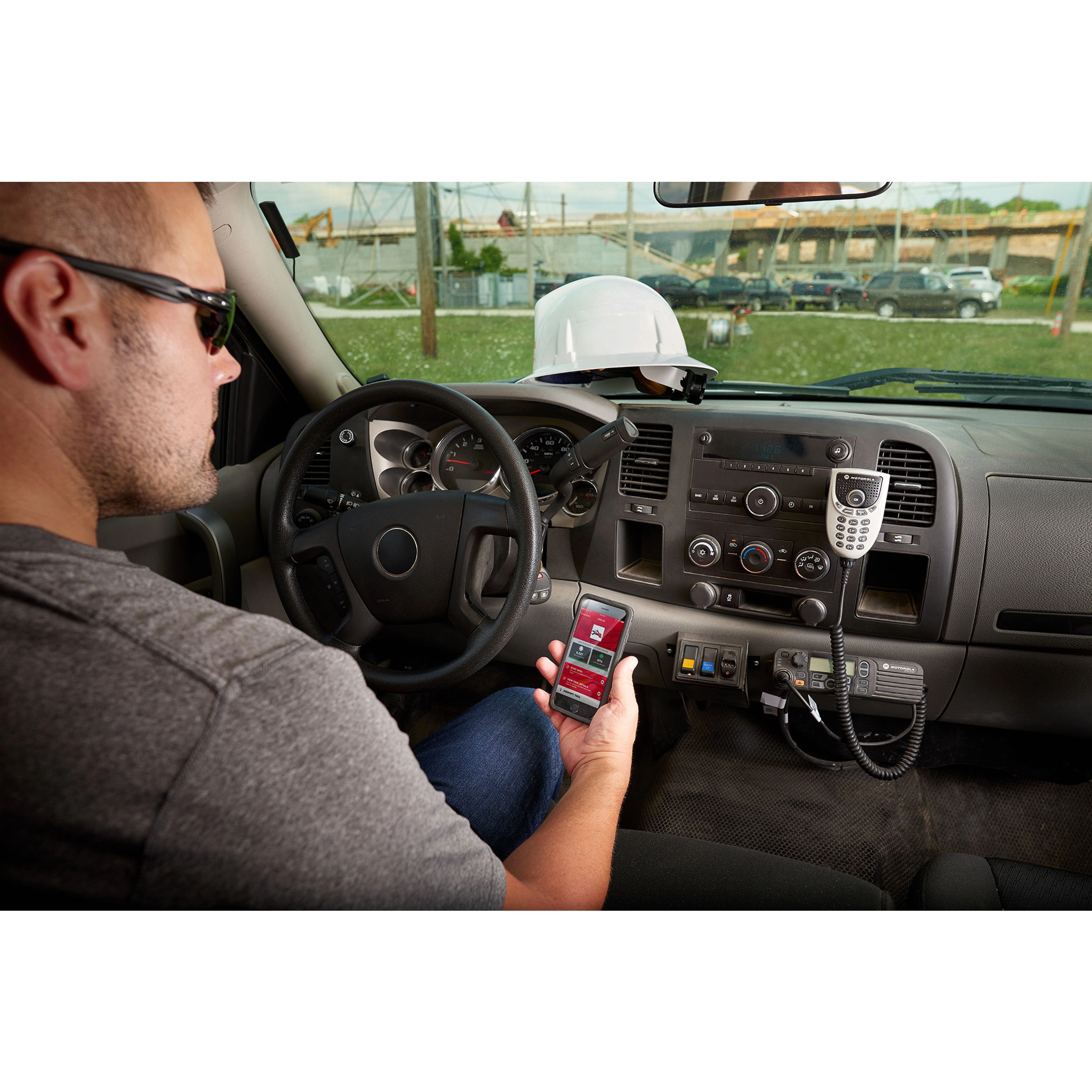 A person is sitting in a vehicle using a wireless remote control for the FORCELOGIC™ M18™ 3" Underground Cable Cutter Kit. The dashboard includes a radio and mounted devices, while a hard hat is placed on the dashboard.