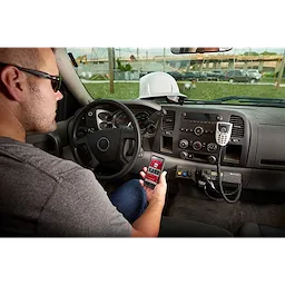 A person is sitting in a vehicle using a wireless remote control for the FORCELOGIC™ M18™ 3" Underground Cable Cutter Kit. The dashboard includes a radio and mounted devices, while a hard hat is placed on the dashboard.
