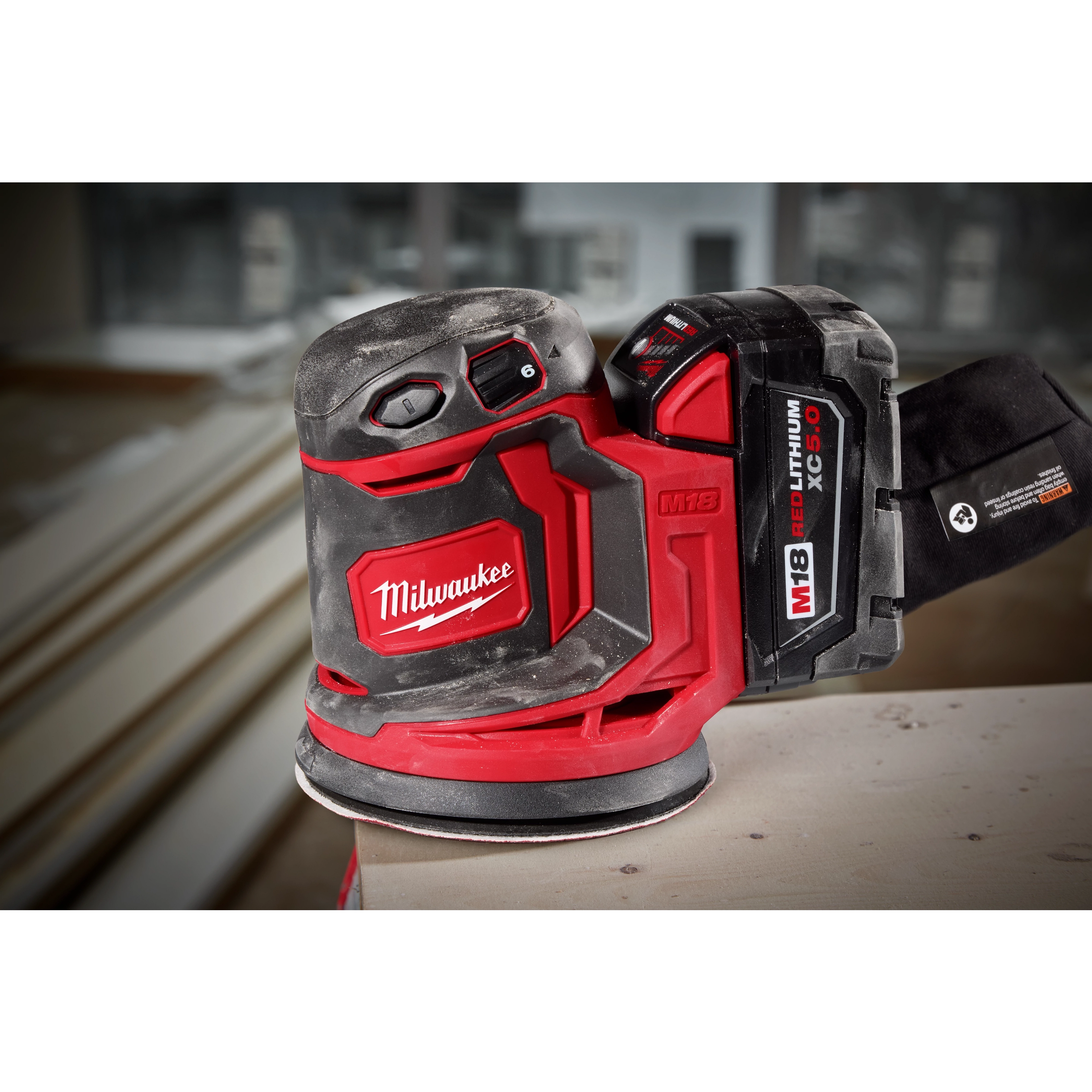 The Milwaukee M18 Random Orbit Sander features ergonomic design with red and black casing, a dust collection system, and a battery pack labeled "M18 REDLITHIUM XC5.0." It is placed on a wooden surface, with building materials visible in the blurred background.