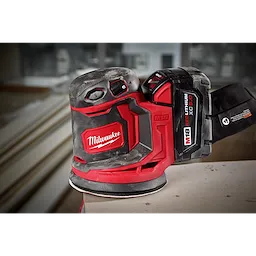 The Milwaukee M18 Random Orbit Sander features ergonomic design with red and black casing, a dust collection system, and a battery pack labeled "M18 REDLITHIUM XC5.0." It is placed on a wooden surface, with building materials visible in the blurred background.
