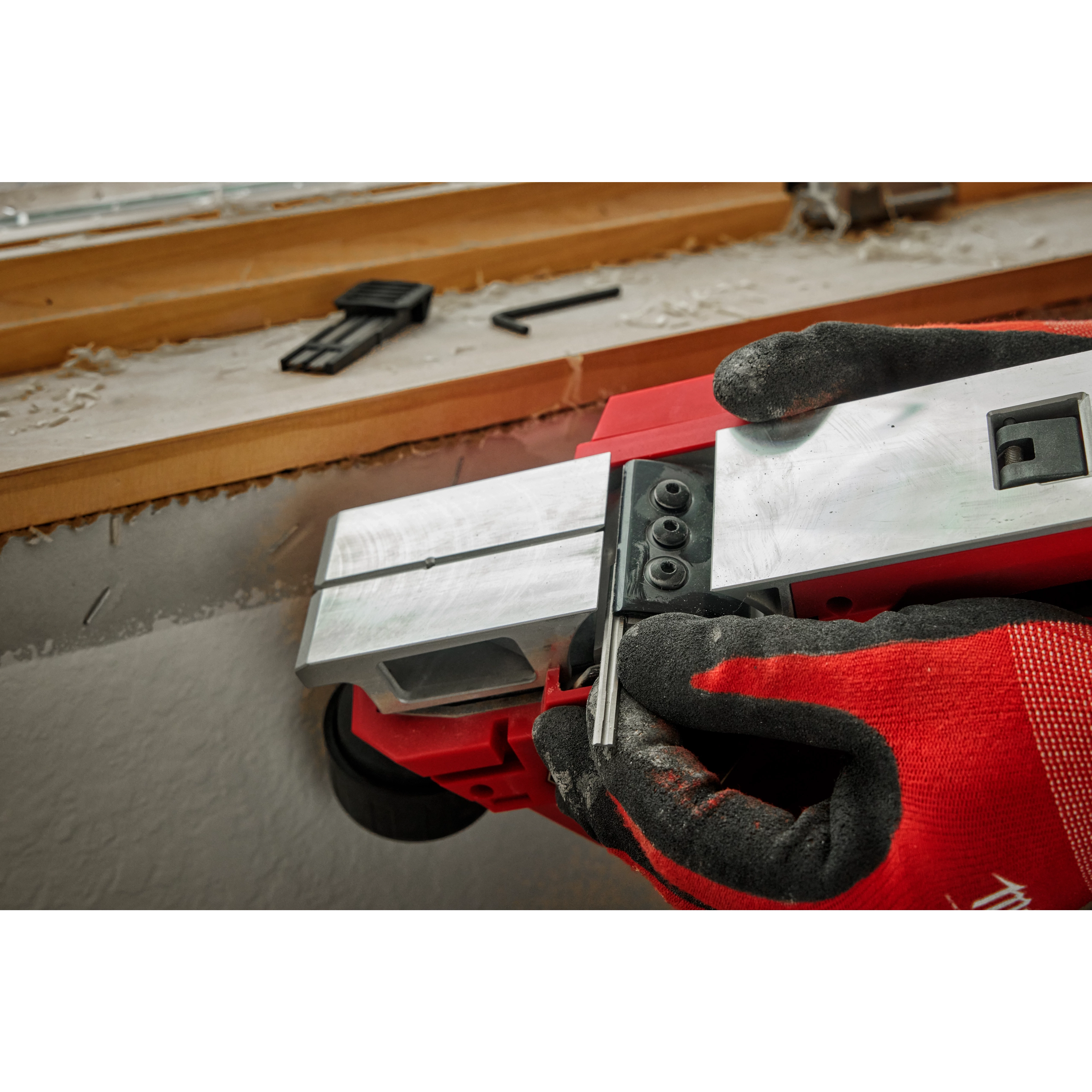 Person adjusting the blade of an M12 Brushless 2-inch Planer with red and black gloves, set on a wooden windowsill.