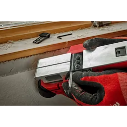 Person adjusting the blade of an M12 Brushless 2-inch Planer with red and black gloves, set on a wooden windowsill.