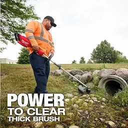 Worker using an M18 FUEL QUIK-LOK String Trimmer Attachment to cut grass near rocks and a drainage pipe.