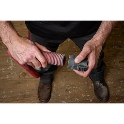 A person holding a striped red and black flexible hose and a cleaning tool from the PACKOUT™ Cleaning Accessory Kit. The person stands on a wooden floor with specks of debris. The focus is on the hands and tools.