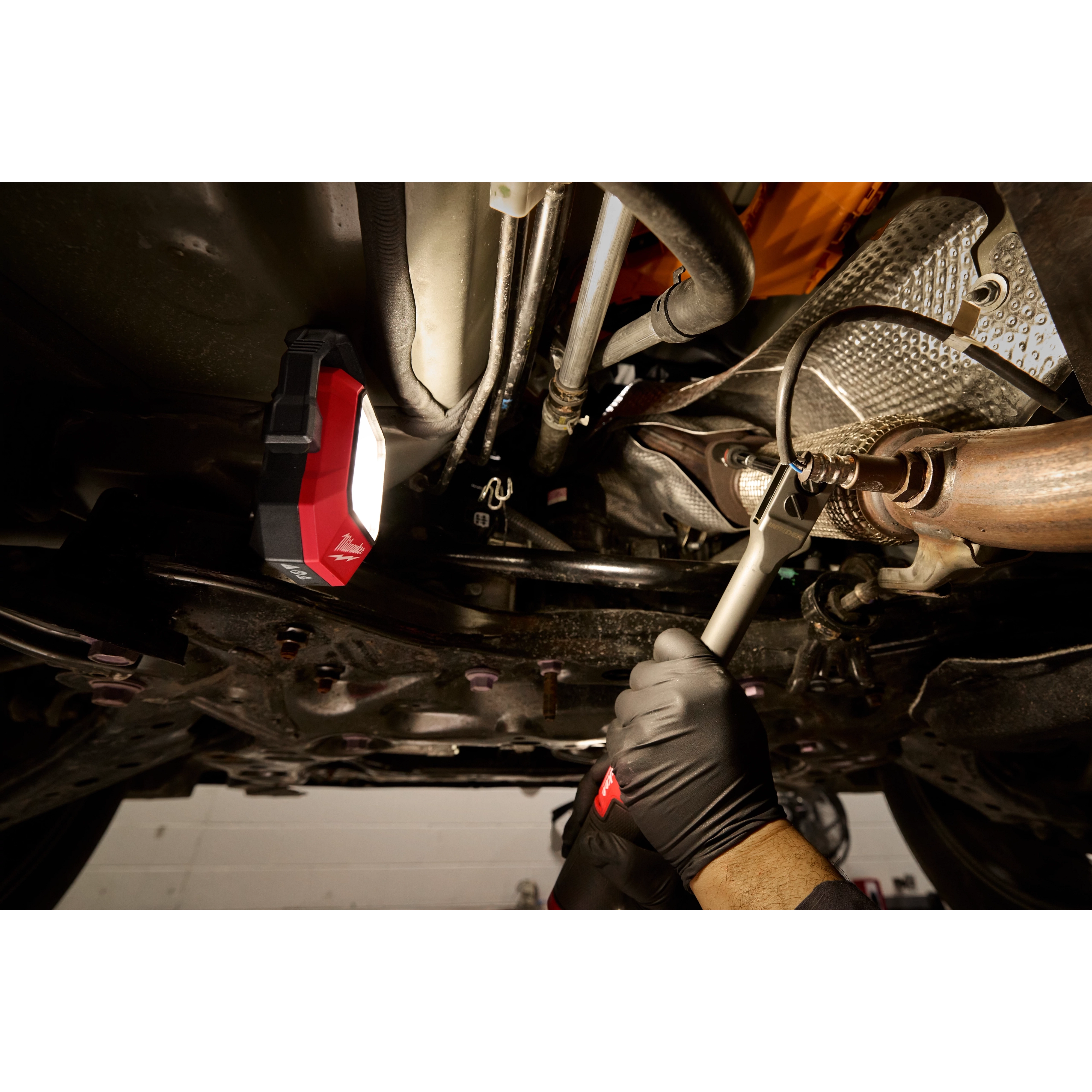 Milwaukee Rechargeable Pivoting Task Light with Spot Mode illuminating the underside of a car while a person uses a wrench.