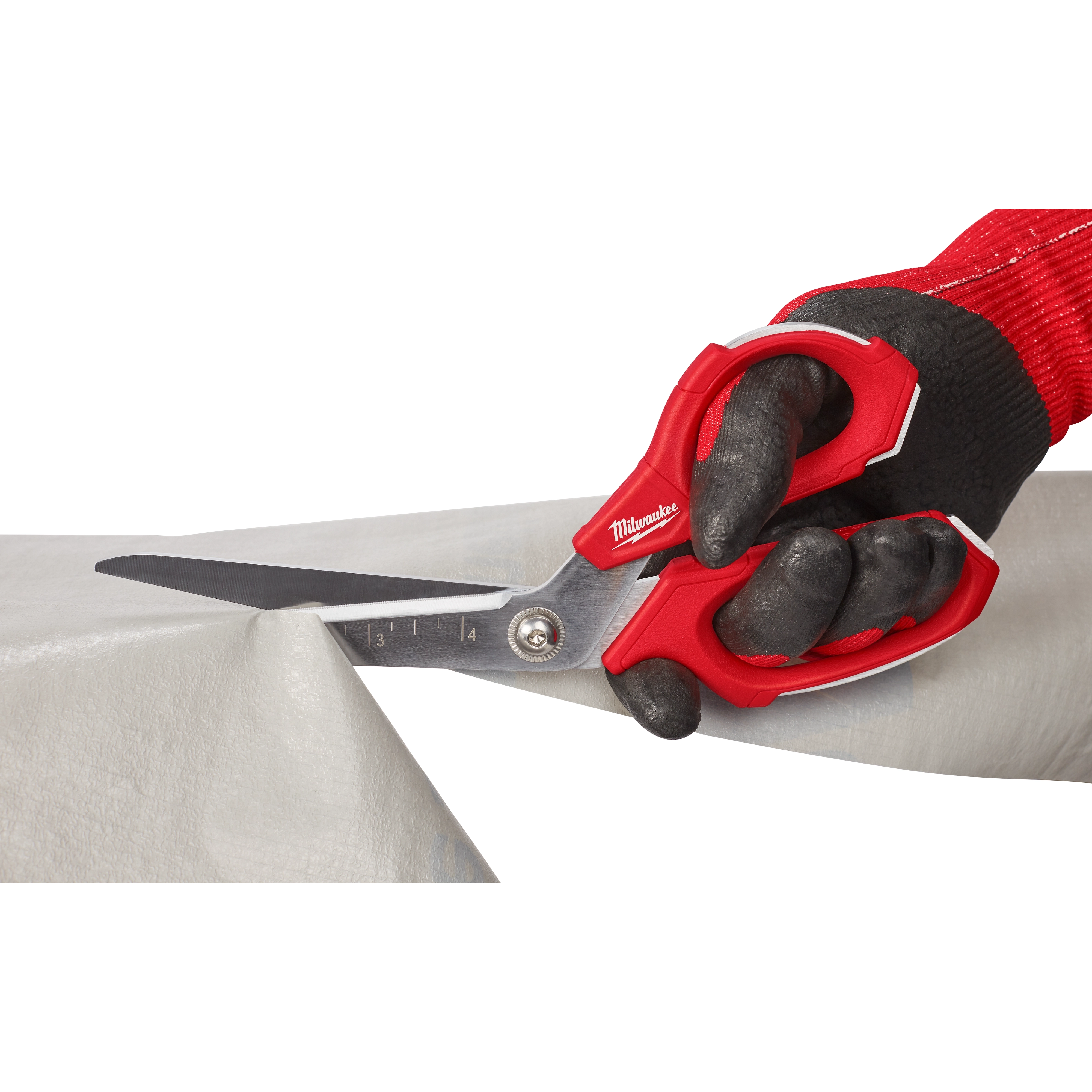 A person wearing black and red work gloves uses Offset Jobsite Scissors to cut through a sheet of material. The scissors have a red and silver design with measurement markings on one blade.