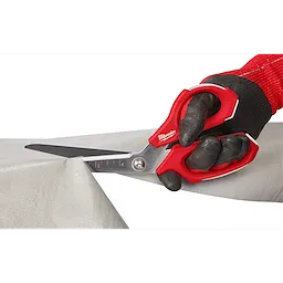 A person wearing black and red work gloves uses Offset Jobsite Scissors to cut through a sheet of material. The scissors have a red and silver design with measurement markings on one blade.