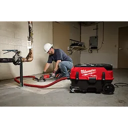 A person wearing a hard hat uses the M18 FUEL™ PACKOUT™ 9 Gallon Dual-Battery Dust Extractor with VACLINK™ in a workshop. The red and black dust extractor is connected to a tool via a red hose. The workplace has concrete flooring and visible pipes.